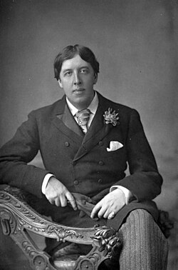 Early middle aged white man in Victorian morning dress, seated with legs crossed, holding gloves and looking pensively towards the camera