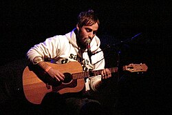 A man in a hoodie playing an acoustic guitar; he is simultaneously singing with a microphone.