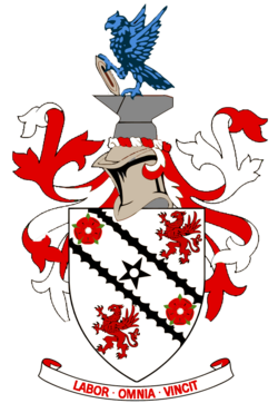 A white shield upon which emblems of two red roses and two red griffins appear at alternate corners. A black five-pointed star is in the centre of the shield's design between two black diagonal lines. Around the shield are red and white ribbons in a symmetrical design. Above the shield is a silver-coloured knight's helmet surmounted by grey anvil upon which a vivid azure-coloured eagle is perched, holding a grey shuttle. Below the shield is the motto "LABOR OMNIA VINCIT".