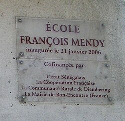 A clear plaque on a grey wall stating "École François Mendy" at the top with smaller writing below