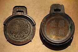 Two circular bronze plates with square handles. The plate on the right is thicker, has four characters embossed into it, and contains a rim around about three-fourths of the edge of the plate. The plate on the left is thinner, and contains a rim that, when the two plates were stacked on one another, would perfectly meet the edges of the first plate.