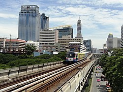 An elevated train, painted in blue, white and a red stripe and with advertisements, running above a road lined with many tall buildings and with many cars