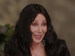 Cher smiles during an interview, wearing a black outfit, long dark wavy hair and statement earrings.