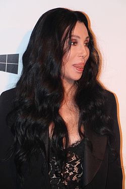 Cher, in a black lace top and blazer, with long, wavy dark hair, looks to the side with her tongue slightly out.