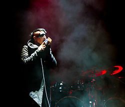 Image of a partially-shaved head, black-haired male performer. He is looking stage left at a 90-degree angle of the camera, near the center of the stage. He is singing in to a microphone while wearing a long, charcoal-black colored pea-coat, with a blue strip of make-up spanning across his face, covering both eyes. Behind him is a drum kit, and the stage is partially obscured by smoke.