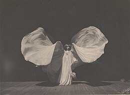 A black and white image of a woman on a stage wearing a dress of long white fabric, which billows out dramatically.
