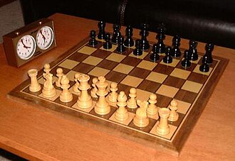 This photo shows a chessboard with pieces set up on both sides, ready to play. A chess clock is at the side.