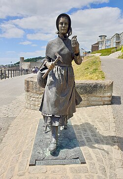 A bronze statue of Mary Anning striding forward, with a pickaxe in one hand and a fossil in the other.