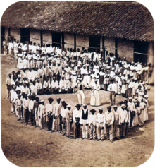 Photograph showing a group of people dressed in white, who have gathered in front of a tile-roofed farm building and observe another large group which has formed a large circle surrounding five men straddling large drums, a woman and two other men