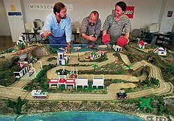 A miniature model of an island is positioned on a table inside an office building, with computers in the background and signs that says "Mindscape" and "Lego" on the wall. Various Lego buildings, vehicles, plants, minifigures, and other pieces are placed on different parts of the island. Three men, development team members Scott Anderson, Wes Jenkins, and Dennis Goodrow, are standing over the model and playing with small Lego vehicles in their hands.