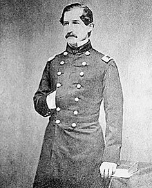 A three-quarters length monochrome photograph of a white man in a dark military uniform; he has dark hair and mustache, his right hand is tucked into the breast of his uniform, and he is looking to the camera's left.