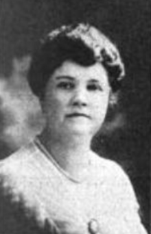 A white woman with coiffed hair, wearing a light-colored lacy blouse with a brooch pinned to the front, and a strand of beads or pearls