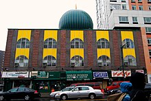 A two-story brick building on a city street with arched windows, a green dome atop in the center and storefronts at street level