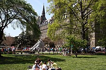 Many students mingle in the background while a group sit in the foreground on a grass lawn. The large stone clocktower is seen above the trees on the lawn.