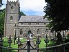 Gray stone building with square tower at left hand end. Foreground includes grass area with gravestones, taken over the top of metal railings.