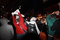 On the crowded sidewalk of a street at night, a person in a red jacket attempts to hold up a sign, and a woman in a black jacket and wearing a gray scarf and hat reaches up to grab the sign.