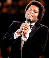 A dark-skinned man wearing a tuxedo, singing into a microphone