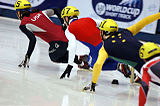 A tight group of four skaters leaning inwards as they make a turn. The four skaters are wearing yellow helmets and suits that the display flag colors of their respective countries. The skaters have their left hands touching the ice for balance as they accelerate around the turn.