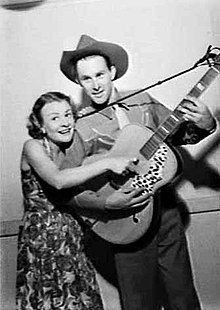 A three-quarter black-and-white image of a couple. The woman at left is wearing a floral dress and has her right hand placed on the string of the guitar. It is being held by the twenty-five-year-old man who wears a cowboy shirt and a hat. His left hand is on the fret board and his right is below the woman's hand.