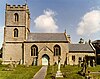 Stone building with square tower at left hand end. In the foreground either side of a path are gravestones in a grassy area.