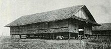 Black-and-white image a grass nipa hut raised a few feet off the ground by wooden supports. Another hut can be seen in the background.