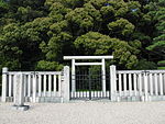 White torii gate behind a concrete fence in front of trees.