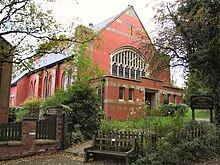 An orange-brick church perched on a shallow hillside in a daytime scene in which the sky appears white. The triangular stone windows of the two-storey church are clear, as are the grounds in the church's proximity. The grounds are composed of green grass, light grey stone pathways, brown brick fencing and green trees overhanging the church.