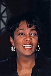 A woman sings while she is recharged on a piano. She wears a long black ensemble and diamond earrings.