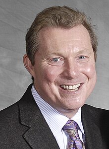 close-up of Jim Donovan wearing a colorful patterned tie, white, striped shirt, and dark blazer, grinning directly at camera