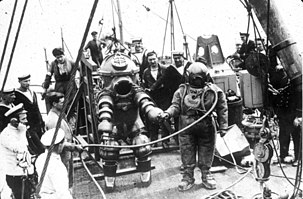 Monochrome view of the deck of a ship with a pair of divers, and a group of onlookers, some of which are crew of the ship. To the left is a diver in an armoured suit, and to the right the diver is in standard diving dress with copper helmet