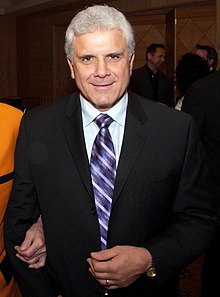 An Italian-Canadian man in his sixties with grey hair, wearing a tuxedo with a light blue shirt and purple tie, facing the camera.