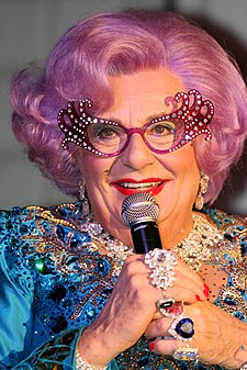 closeup of Dame Edna wearing sparkly blue dress, over-the-top eyeglass frames, and multiple finger rings