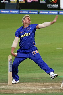 A white coloured man in his bowling action. He is wearing a blue outfit and is alongside the cricket stumps. The boundary ropes can be seen in the background.