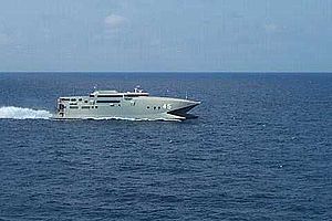 A warship-grey catamaran travelling at speed (from right to left) on the open sea