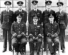 Portrait of eight men in dark military uniforms with peaked caps, five standing and three seated