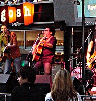 A band is performing outside a shop forefront. The male at left is playing a guitar and is at a microphone. A fifty-seven year-old male in middle is shown partly in left profile. He has dark hair, wears sunglasses and is playing a guitar while at a microphone. A drum kit is to his left and behind, but the drummer is not in shot. A third band member, also on guitar, is mostly obscured by frame cut-off. Two audience members are in the foreground.