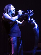 A man with his eyes closed and mouth open, holding a microphone; he is wearing dark clothing and wrist bands.