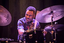 Nate Smith playing the drums on in front of a dark curtain