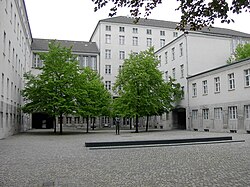 View of a courtyard from one of its sides; the courtyard is laid with brick and spotted with small trees. The courtyard is surrounded by white and gray buildings that range from two stories to six stories.