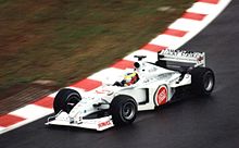 A figure, wearing a helmet of a dark blue design with red and yellow stripes, is driving a Formula One with an white colour scheme adorned with sponsorship. He is holding both hands on a steering wheel, turning right.