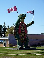 A large turtle statue standing on two legs and holding a Canadian flag in one hand an American flag in the other.