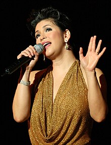 A woman in a gold dress sings into a hand-held microphone.