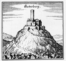 A medieval fortress stands on the peak of a hill, dominating the valley below it, and the river passing it in the distance; the fortress has a central keep (tower), and an elaborate ring of crenelated walls and narrow windows. A road twists around the mountain to the gate.