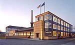 A factury building in modernist style with a chimney and two flags. Evening photo.