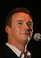 A close angle image of Watson in a black suit and smiling during a performance with a microphone mounted on a stand