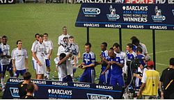 A coloured photograph of the Chelsea squad standing on a podium, celebrating their second Premier League Asia Trophy win.