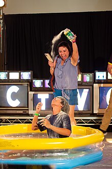 Thach's mother pouring Parmesan cheese over Thach's head, who is sitting in an inflatable pool, looking up