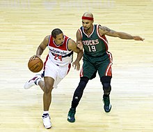 At left man carrying the ball wearing a white Wizards uniform, man at right guarding him wearing a red headband and dark green Bucks uniform