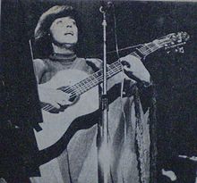 Black and white photograph of a short-haired woman in a poncho standing behind a microphone, playing a guitar.