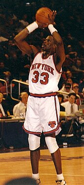 An African American basketball player attempts a jump shot. He wears a white jersey with an orange "NEW YORK" and "33", and spectators can be seen in the background.